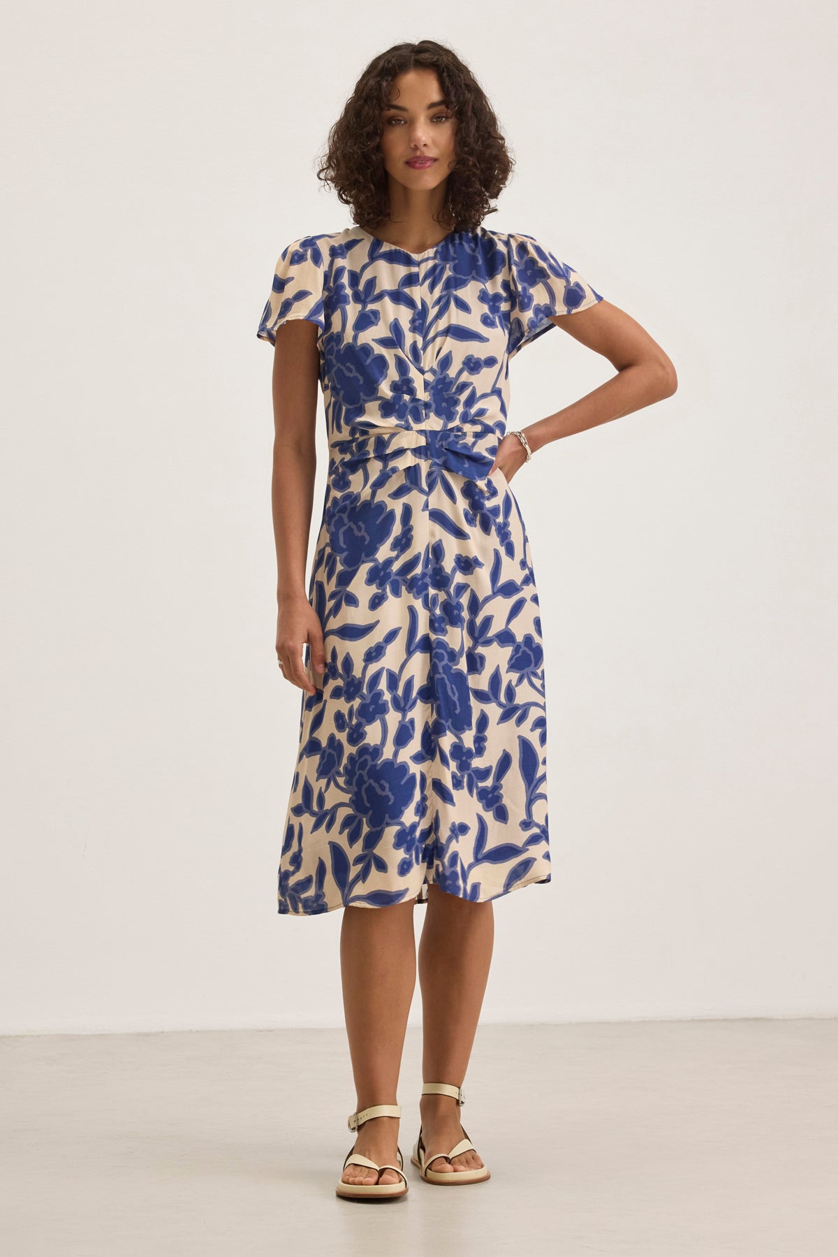 A woman models the NEVINE DRESS from Velvet by Graham & Spencer, featuring a beige base with blue floral patterns, paired with beige sandals against a plain backdrop.-38918753452225