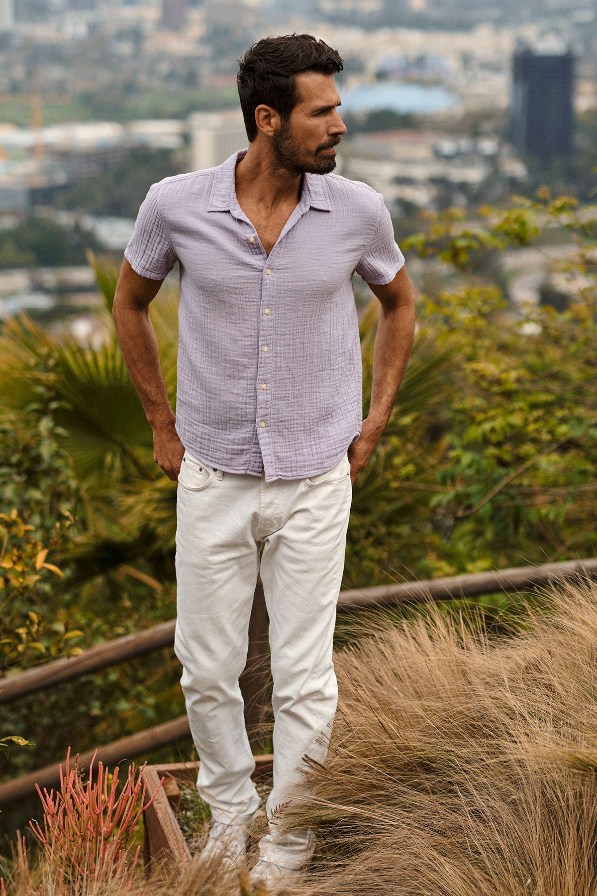  A man in Velvet by Graham & Spencer white cotton gauze pants standing Christian Button-Up Shirt on a hill overlooking the city. 