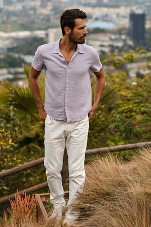 A man in Velvet by Graham & Spencer white cotton gauze pants standing Christian Button-Up Shirt on a hill overlooking the city.