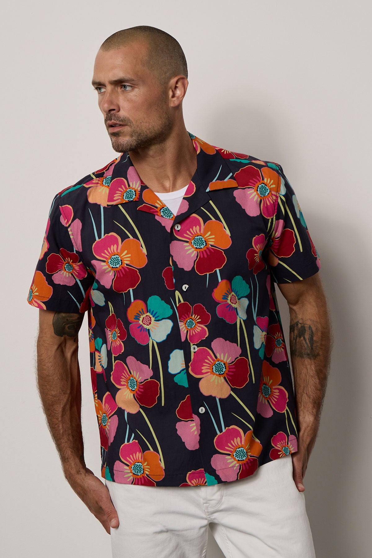 A man wearing a Velvet by Graham & Spencer Iggy printed button-up shirt with a bold floral print and white pants, perfect for summer vacations.-35678388191425