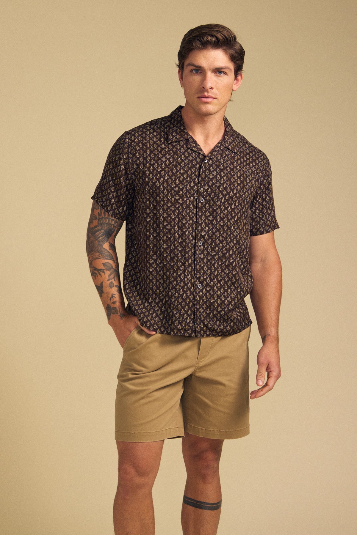 A man stands against a beige background wearing the EMILIO SHIRT by Velvet by Graham & Spencer and tan shorts. He has a tattoo on his left arm and leg.-41701960188097