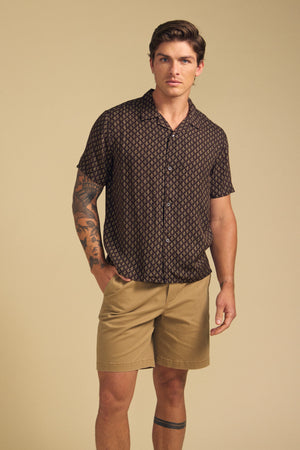 A man stands against a beige background wearing the EMILIO SHIRT by Velvet by Graham & Spencer and tan shorts. He has a tattoo on his left arm and leg.