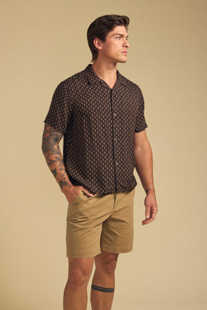 A man stands against a beige background wearing the EMILIO SHIRT by Velvet by Graham & Spencer—a black patterned camp collar button-up paired with tan shorts, adding effortless style to his relaxed look.