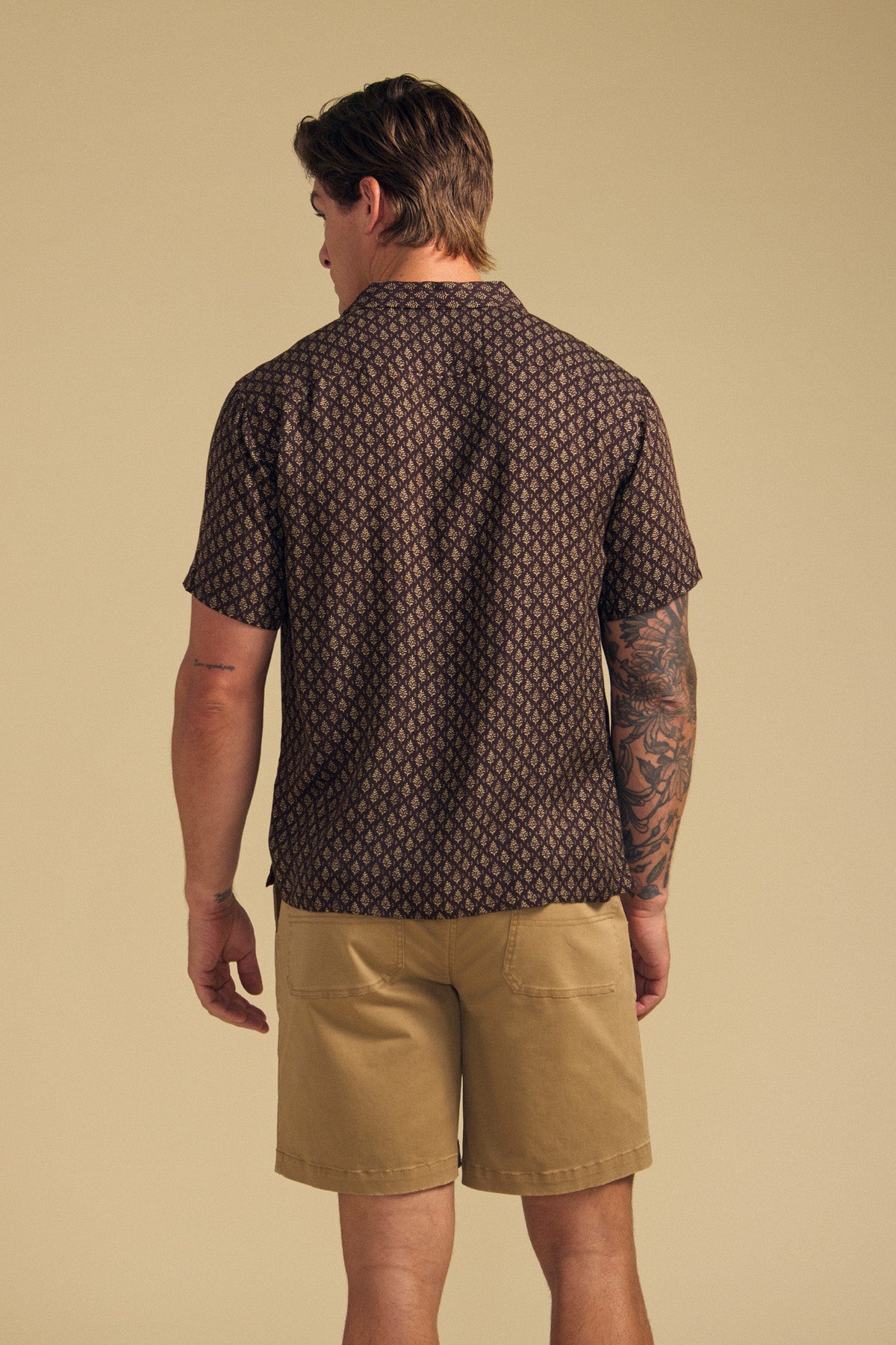 A man faces away, wearing the EMILIO SHIRT by Velvet by Graham & Spencer with a camp collar and patterned design, paired with khaki shorts. A tattoo sleeve is visible on his left arm against a plain beige background.-41701960089793