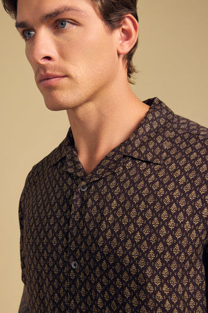 A man wearing the EMILIO SHIRT by Velvet by Graham & Spencer, featuring a brown camp collar and a small beige leaf pattern, stands against a neutral background, looking off to the left.