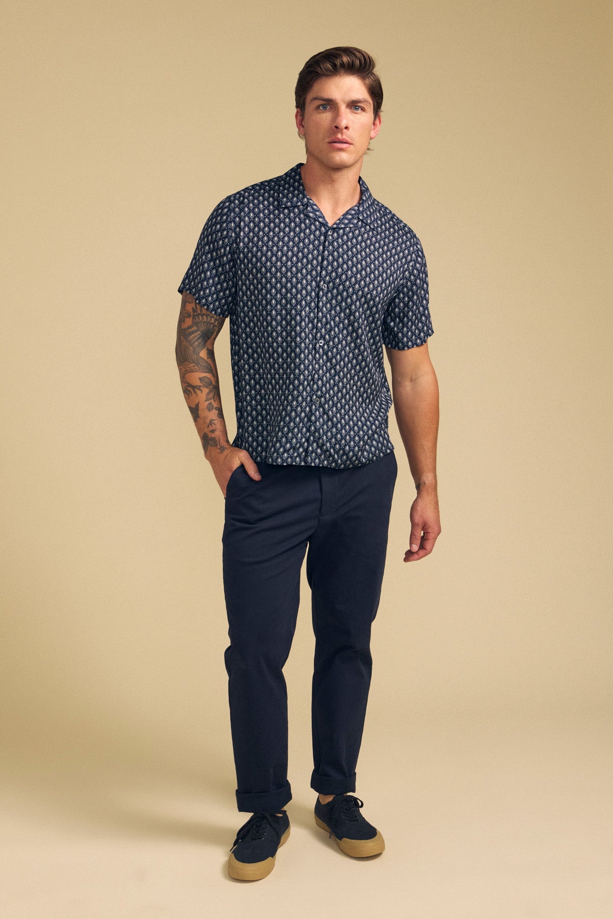 A man stands against a beige background wearing the EMILIO SHIRT by Velvet by Graham & Spencer, paired with dark pants and black sneakers with tan soles. His tattooed right arm adds edge, while the breathable modal fabric ensures cool comfort.