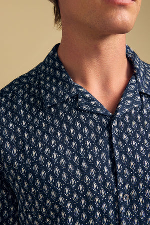 A person wearing the EMILIO SHIRT by Velvet by Graham & Spencer, a relaxed fit navy camp collar shirt with a white geometric leaf pattern, made from breathable modal for all-day comfort. Shown from the shoulders up.