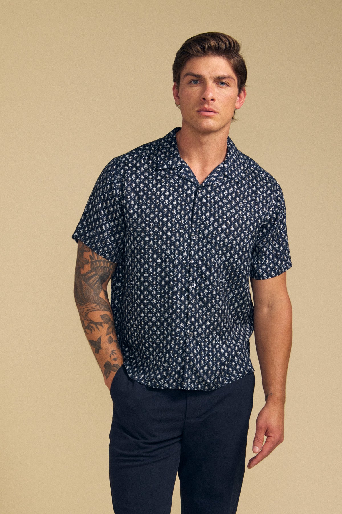 A man with short brown hair wears the EMILIO SHIRT by Velvet by Graham & Spencer—navy camp collar, relaxed fit, and dark pants—standing against a beige background with arm tattoos, exuding effortless laid-back style.
