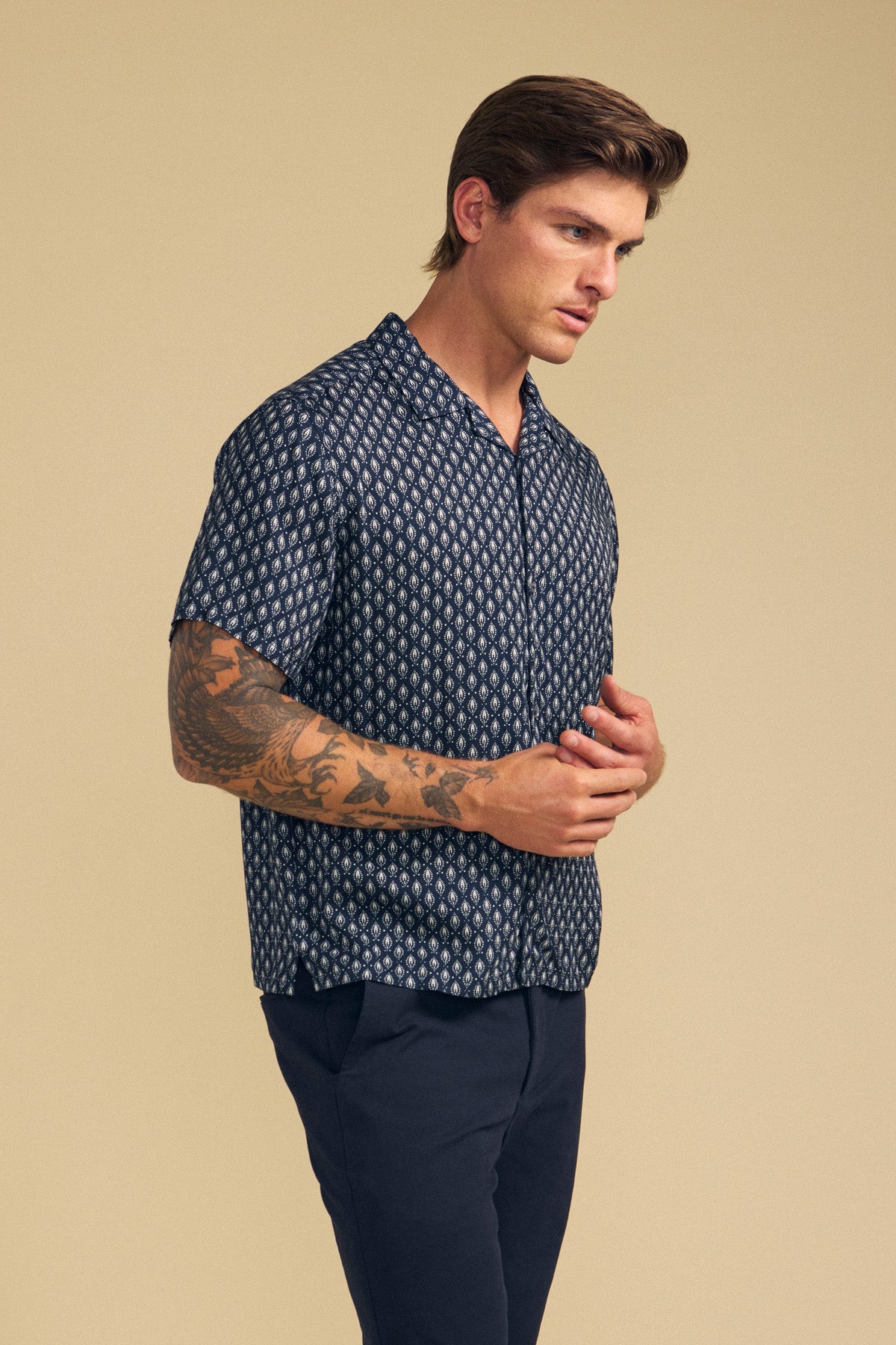 Man with tattoos on his left arm wears the EMILIO SHIRT by Velvet by Graham & Spencer, a relaxed fit button-up camp collar style, paired with dark pants against a beige background.