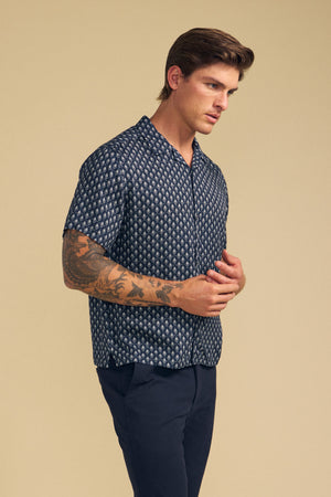 Man with tattoos on his left arm wears the EMILIO SHIRT by Velvet by Graham & Spencer, a relaxed fit button-up camp collar style, paired with dark pants against a beige background.