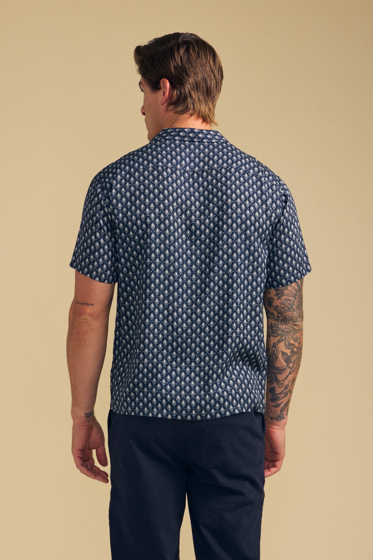 A man faces away, wearing the EMILIO SHIRT by Velvet by Graham & Spencer—a relaxed fit, patterned navy camp collar button-up—paired with dark trousers against a plain beige background.