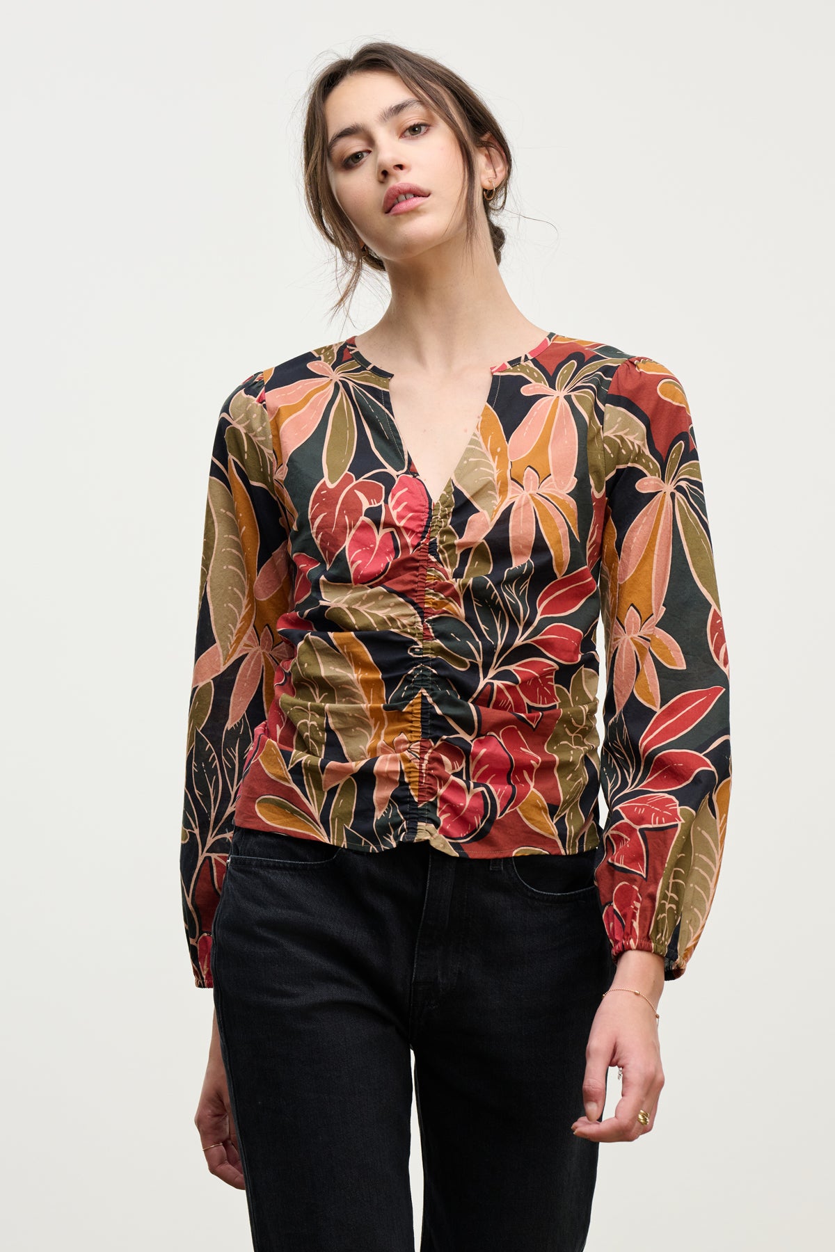 A woman wears the Velvet by Graham & Spencer SYDNEY TOP, a long-sleeve V-neck with a multicolored leaf pattern, paired with black pants, standing against a plain light background.-39742395711681