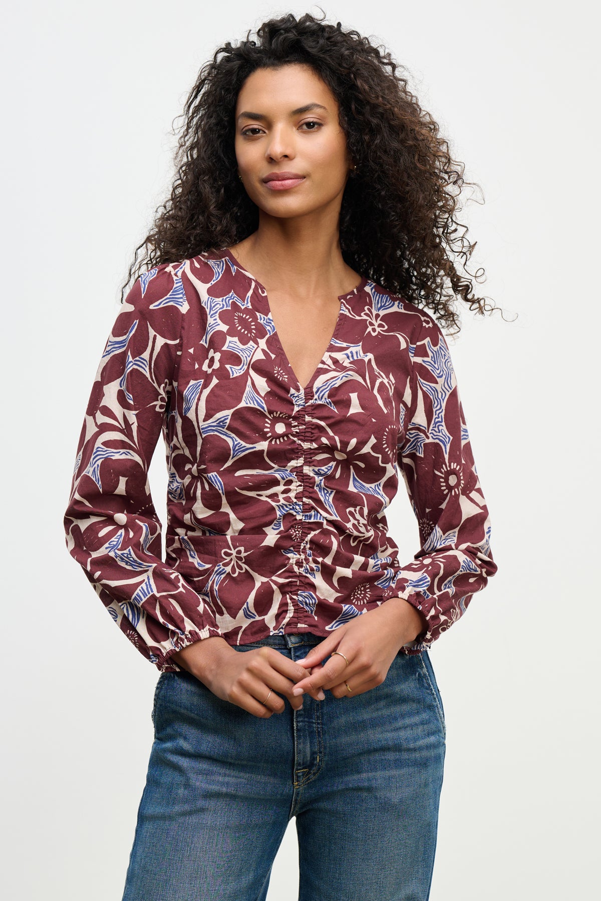 A woman wears the SYDNEY TOP by Velvet by Graham & Spencer—a long-sleeve, ruched, floral cotton cambric blouse in burgundy tones with blue and white patterns—paired with blue jeans, standing against a plain light background.-39617810268353