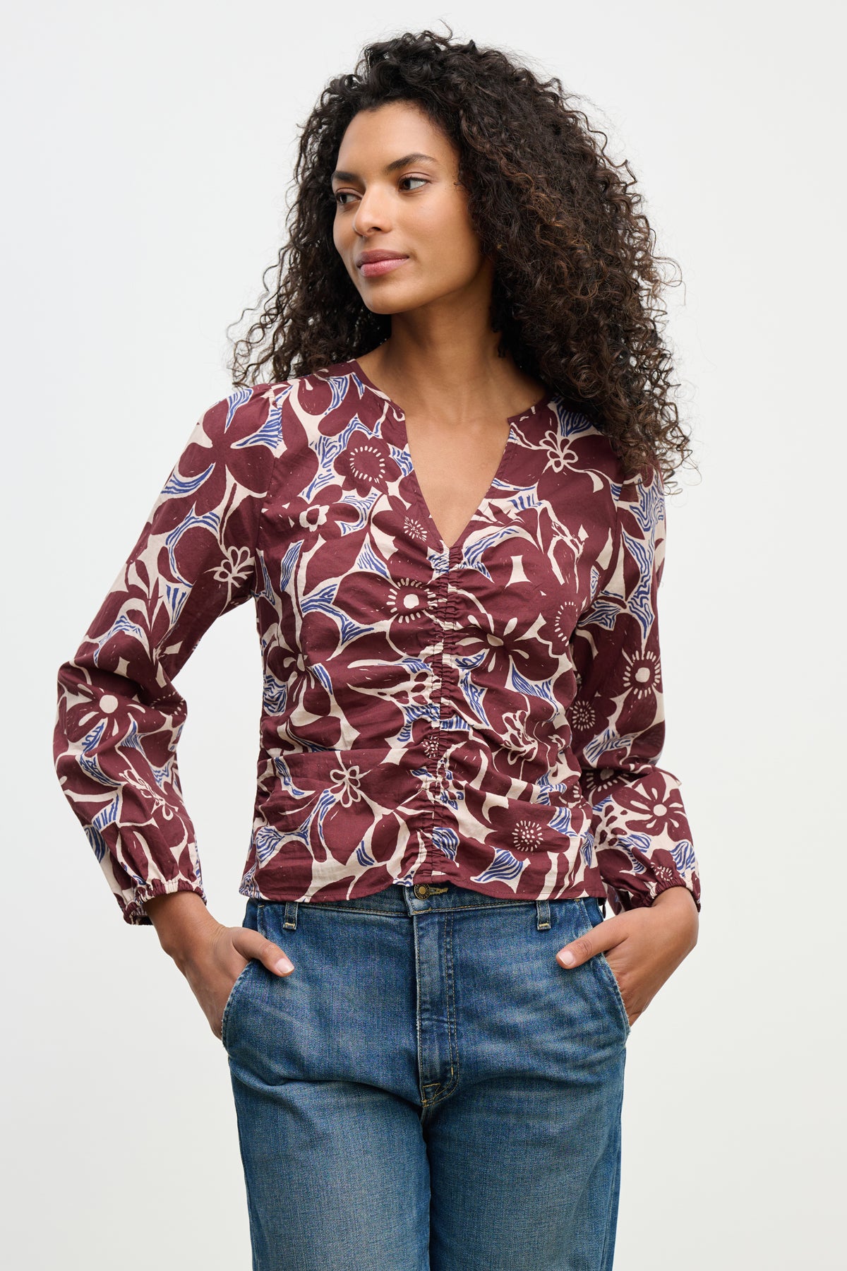 A woman wears the SYDNEY TOP by Velvet by Graham & Spencer, a long-sleeve floral maroon V-neck blouse with blue and white accents, paired with blue jeans, standing with hands in pockets against a plain background.-39617810235585