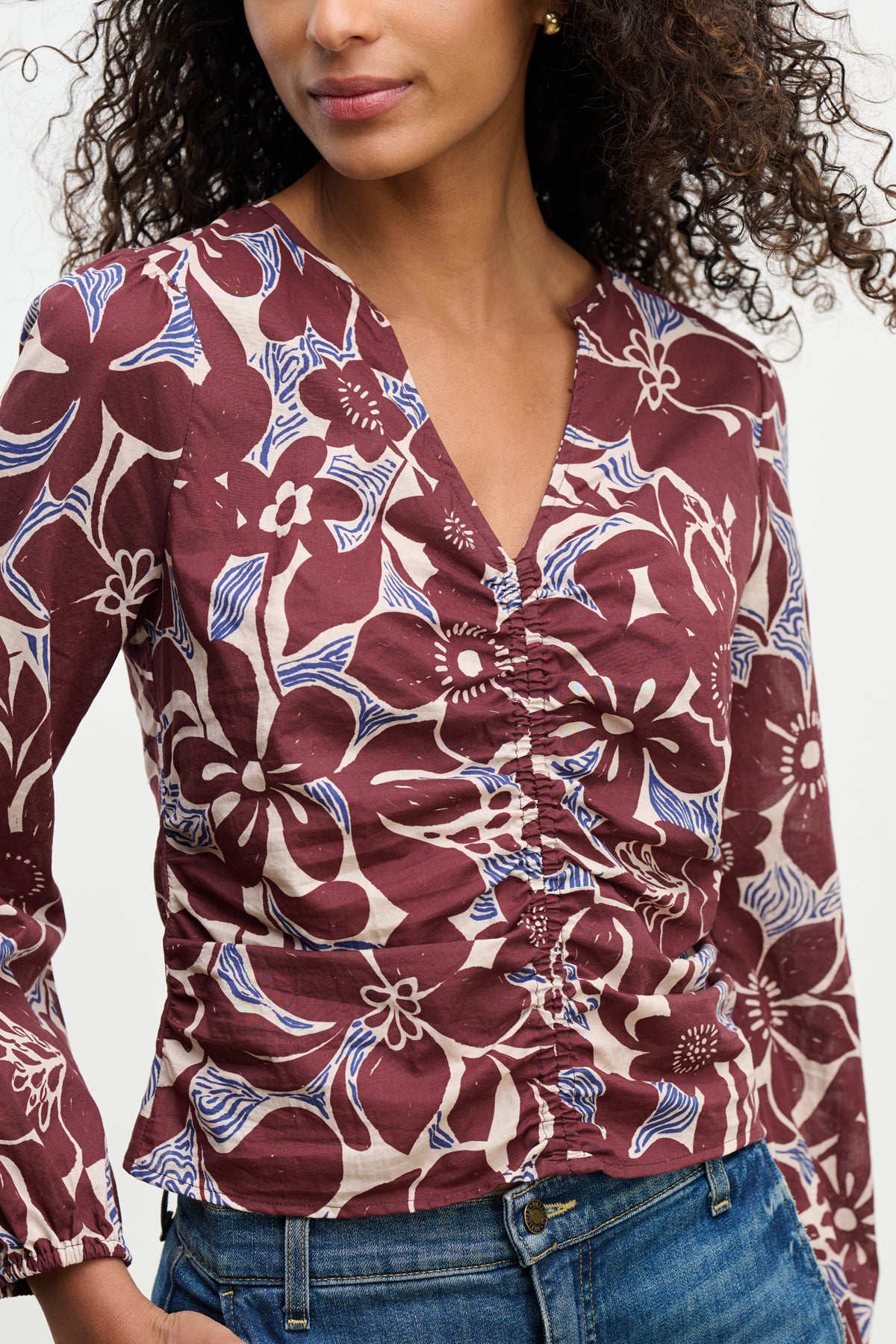 A person wears the SYDNEY TOP by Velvet by Graham & Spencer—a maroon floral-printed cotton cambric blouse with a V-neckline and blue, white accents—paired with blue jeans against a plain background.-39617810104513