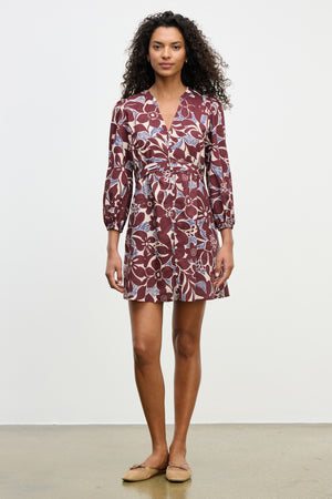 A woman in the TESSA DRESS by Velvet by Graham & Spencer—featuring a maroon V-neckline and white-blue floral pattern—pairs it with tan flats and stands against a plain white background.