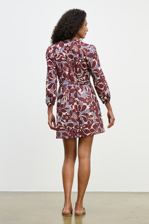 A woman stands facing away, wearing the Velvet by Graham & Spencer TESSA DRESS, a maroon floral pleated mini dress, with sandals against a plain white background.