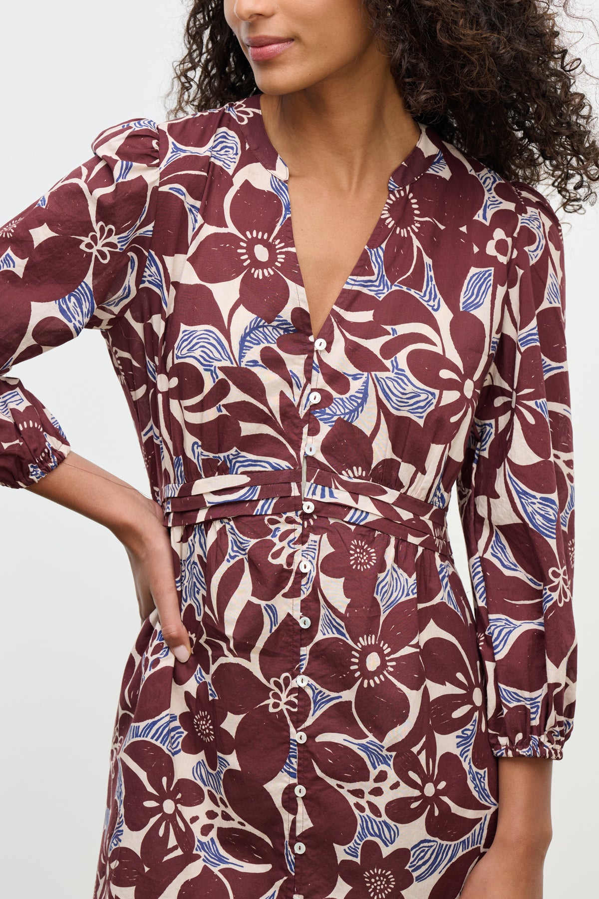 A woman wearing the TESSA DRESS by Velvet by Graham & Spencer, a maroon and beige printed cotton cambric dress with three-quarter sleeves and a V-neckline, stands with one hand on her hip.-39617820950721