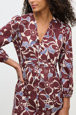 A woman wearing the TESSA DRESS by Velvet by Graham & Spencer, a maroon and beige printed cotton cambric dress with three-quarter sleeves and a V-neckline, stands with one hand on her hip.