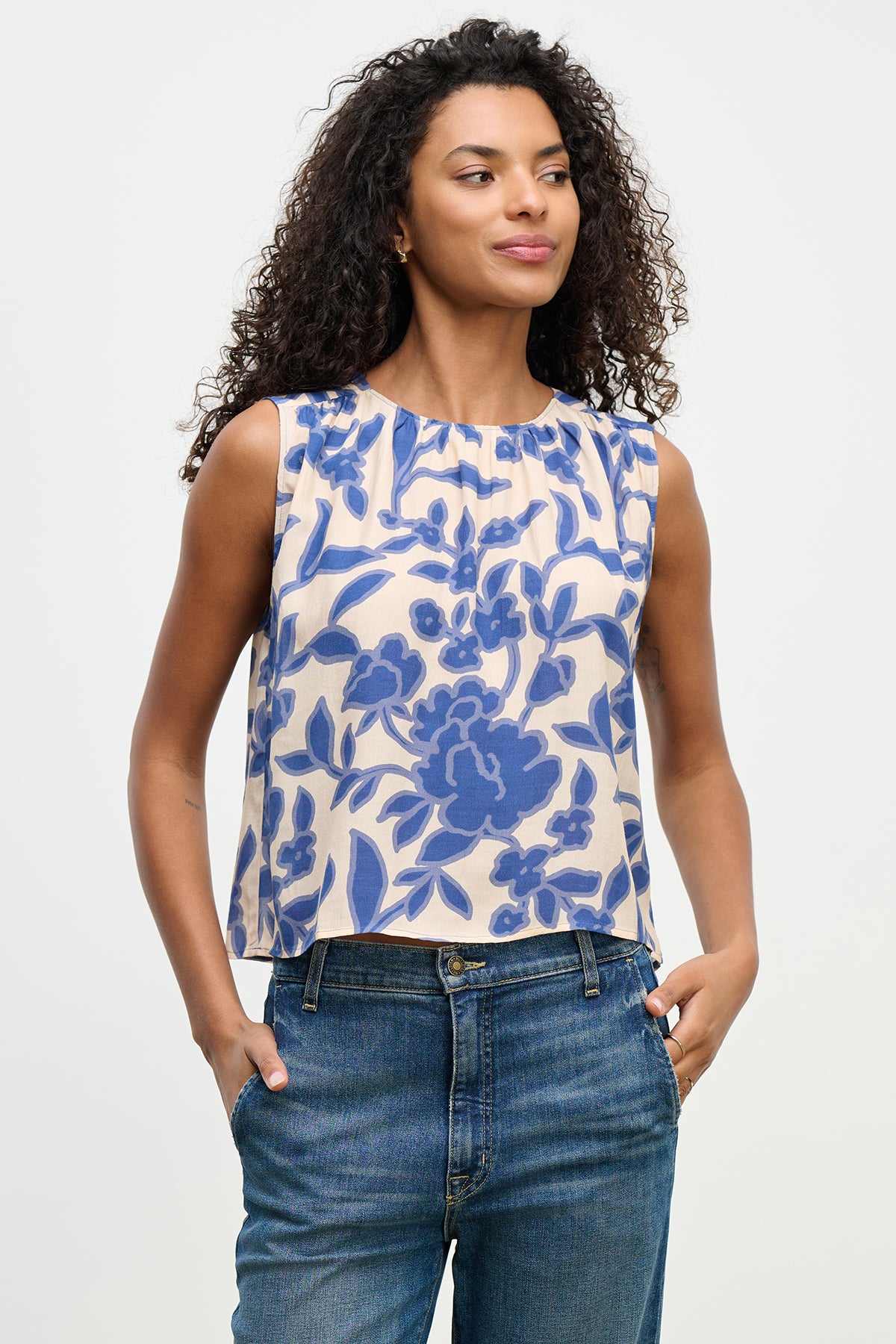 A person confidently poses against a plain background in the KATERINA TOP from Velvet by Graham & Spencer. The sleeveless blue floral top, made from textured viscose royal crepe fabric, adds elegance as they tuck their hands casually into their jeans pockets.-38662830489793