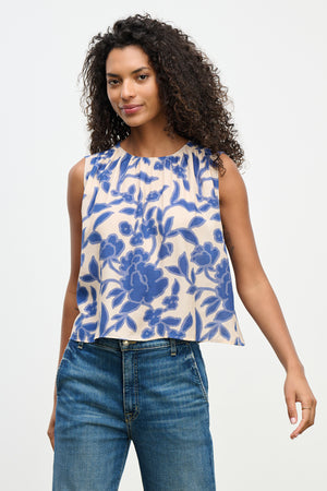 A person with curly hair stands against a plain background, showcasing the Velvet by Graham & Spencer KATERINA TOP, a sleeveless blouse crafted from textured viscose royal crepe with a blue floral pattern.