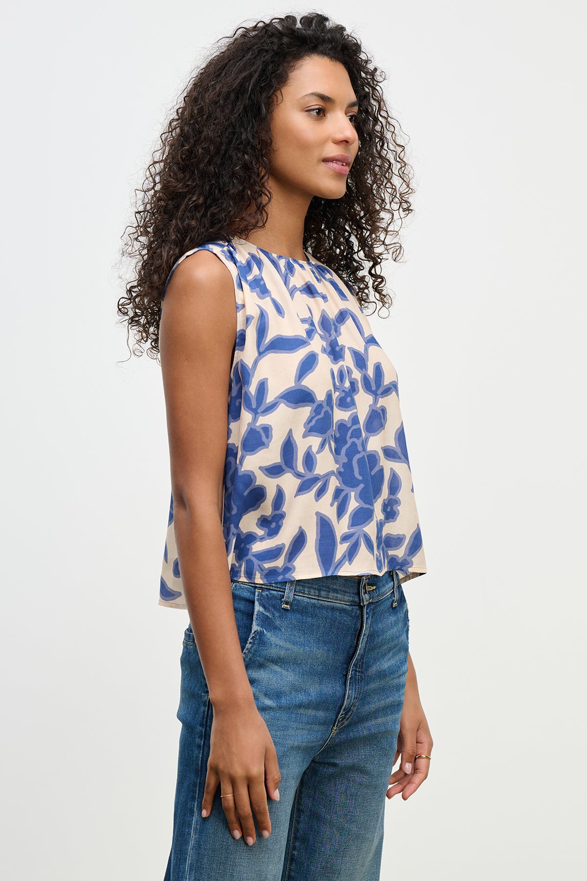 A woman with curly hair in the KATERINA TOP by Velvet by Graham & Spencer, featuring a blue floral pattern and sleeveless design, wears jeans against a plain background.-38662830620865