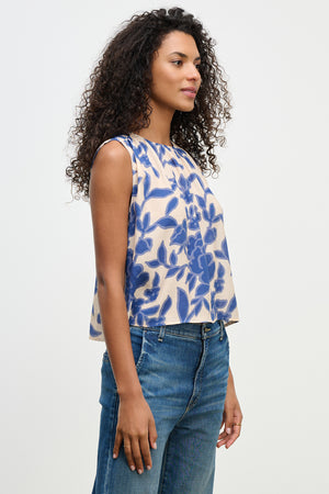 A woman with curly hair in the KATERINA TOP by Velvet by Graham & Spencer, featuring a blue floral pattern and sleeveless design, wears jeans against a plain background.
