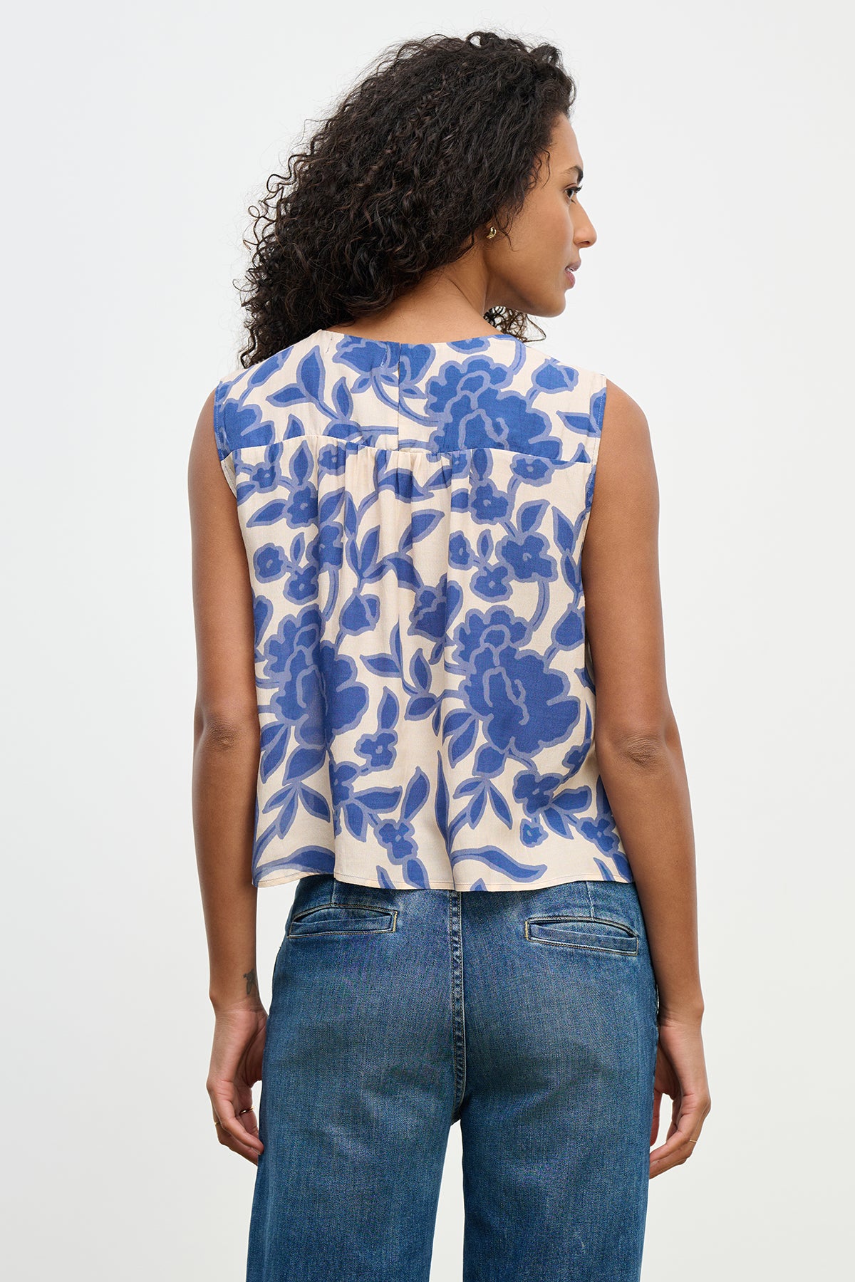 A woman with curly hair, viewed from the back, wears Velvet by Graham & Spencer's KATERINA TOP, a sleeveless design featuring a blue floral pattern, paired with blue jeans.-38662830686401