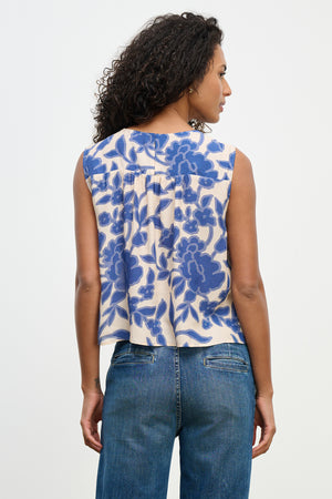 A woman with curly hair, viewed from the back, wears Velvet by Graham & Spencer's KATERINA TOP, a sleeveless design featuring a blue floral pattern, paired with blue jeans.