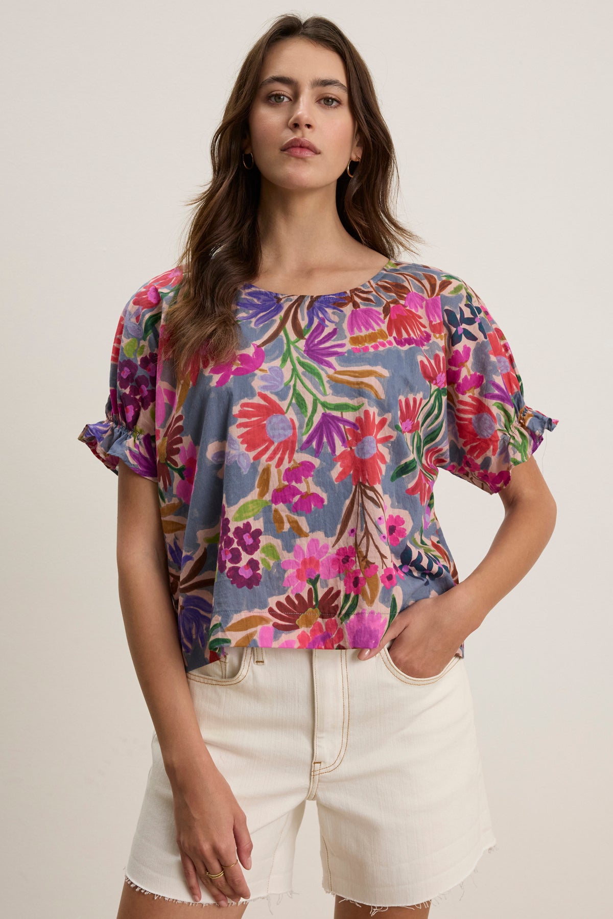 A woman wears the DELILAH BLOUSE by Velvet by Graham & Spencer, a loose, short-sleeve printed top in vibrant colors, paired with ivory high-waisted shorts. She stands facing forward with one hand in her pocket.-39111853211841