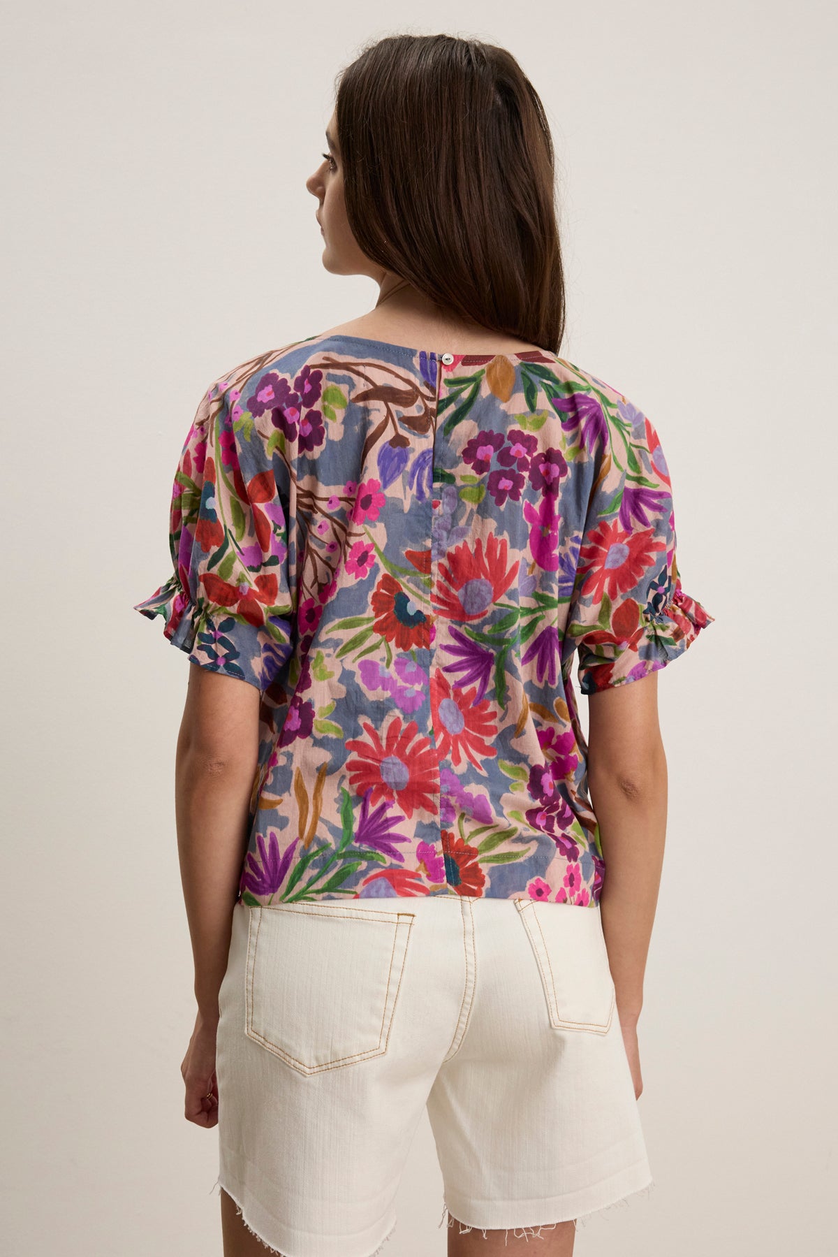 A person stands facing away, wearing the DELILAH BLOUSE by Velvet by Graham & Spencer—a printed soft cotton voile blouse with short dolman sleeves—paired with white high-waisted shorts.-39111853179073