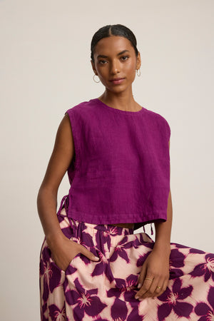 A woman wears the TAVIA LINEN TOP by Velvet by Graham & Spencer with a pink and purple floral skirt, seated against a plain light background.