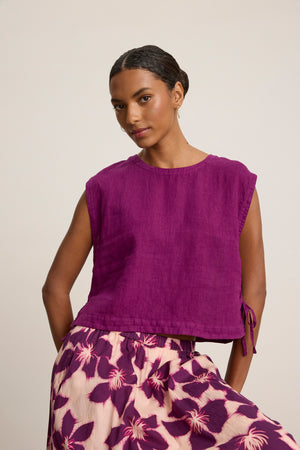 A woman wears the Velvet by Graham & Spencer TAVIA LINEN TOP, a sleeveless purple cropped top, paired with a cream skirt featuring purple floral patterns, standing against a plain background.