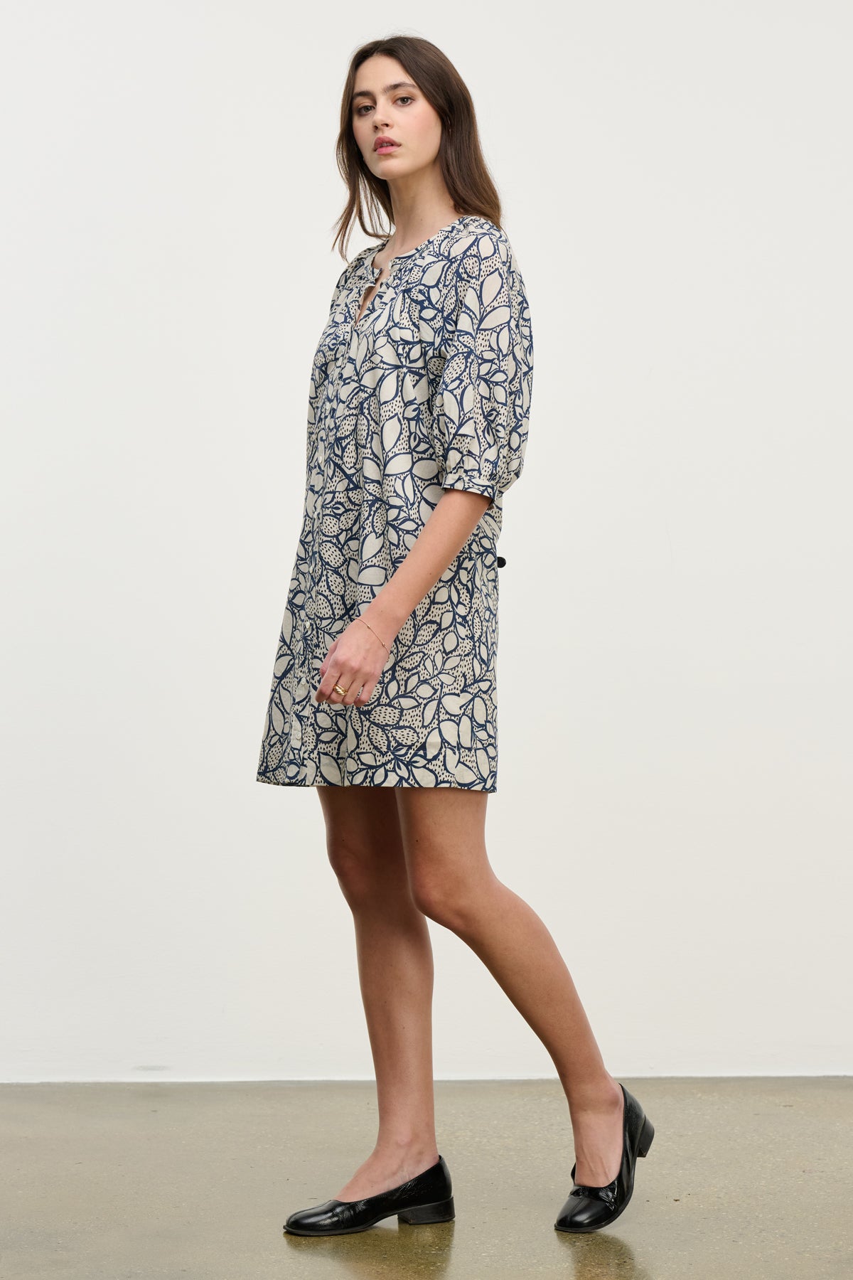 A woman stands sideways against a plain background wearing the SUMMER DRESS by Velvet by Graham & Spencer—a knee-length, patterned cotton voile dress with elbow-length sleeves—paired with black flats.-40227164061889