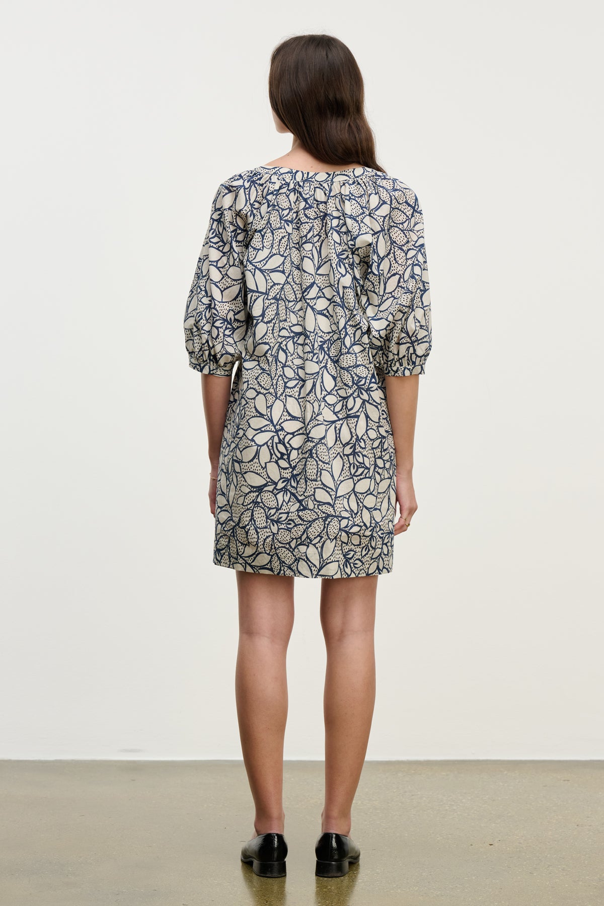 A woman faces away, wearing the SUMMER DRESS by Velvet by Graham & Spencer—a knee-length blue and beige cotton voile dress with elbow-length sleeves—paired with black slip-on shoes against a plain background.