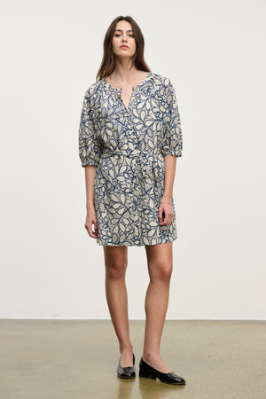 A woman stands against a plain background wearing the SUMMER DRESS by Velvet by Graham & Spencer, featuring a knee-length cotton voile fabric, blue and beige leaf pattern, belted waist, puff sleeves, and black flats.