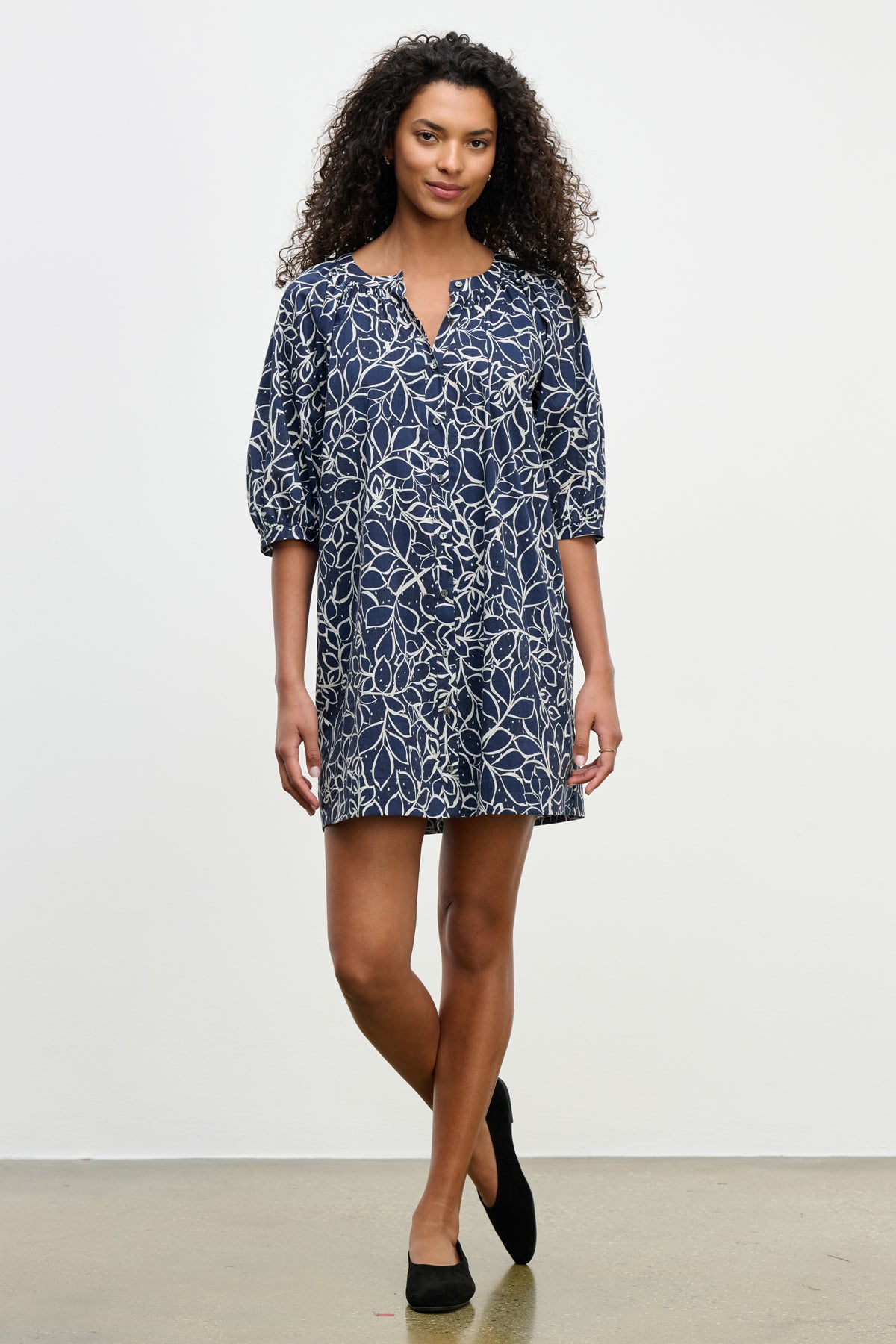 A woman stands against a plain background wearing the SUMMER DRESS by Velvet by Graham & Spencer, a navy cotton voile mini dress with a white floral pattern, puff sleeves, and black flats.-39538953224385