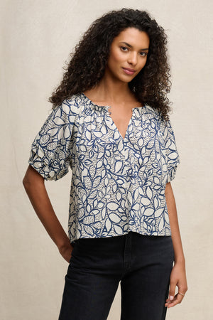 A woman with curly hair wears the THERA TOP by Velvet by Graham & Spencer, a sheer cotton voile blouse with a smocked elastic neckline and blue and white leaf pattern, paired with black jeans against a neutral background.