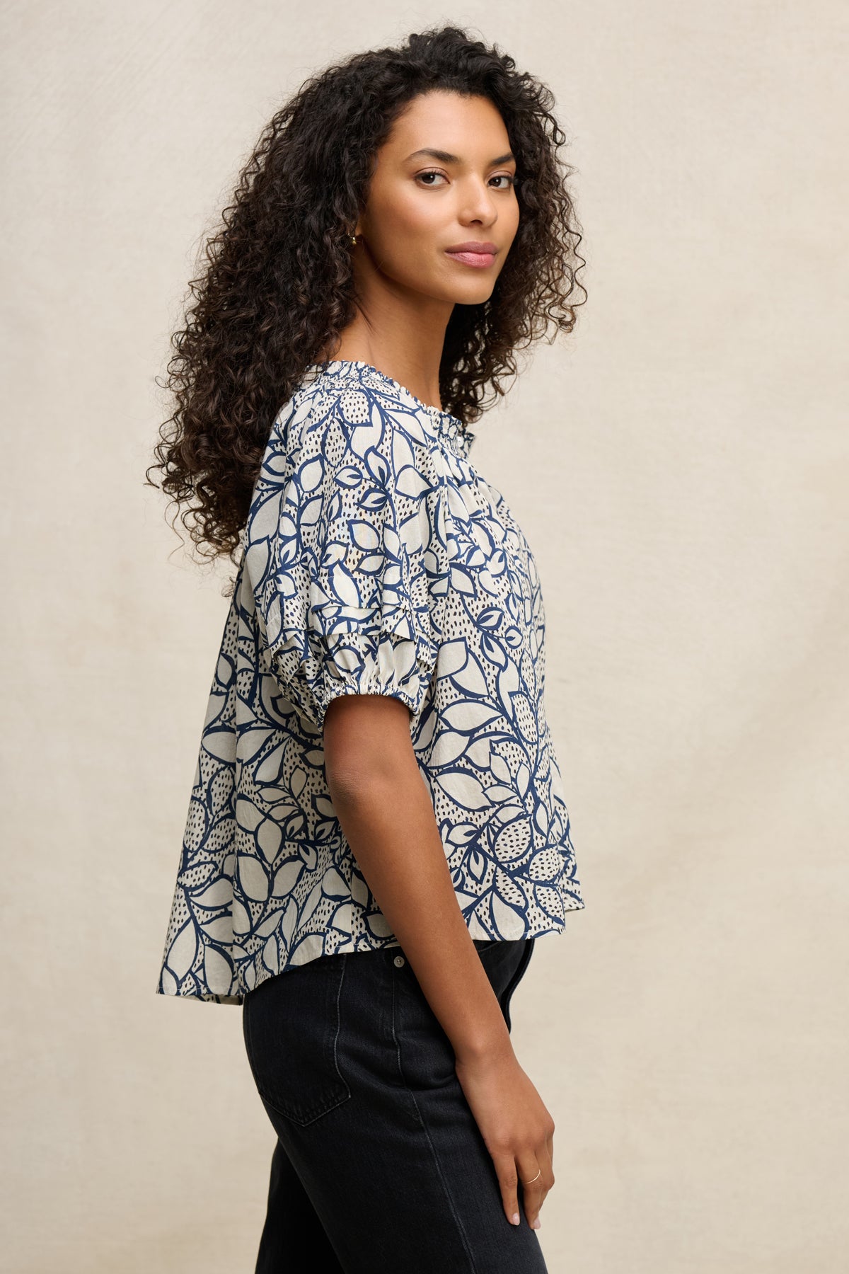 Woman with curly hair stands in profile wearing the THERA TOP by Velvet by Graham & Spencer, a short-sleeve cotton voile blouse with a smocked neckline, paired with dark pants against a neutral background.-39539195052225