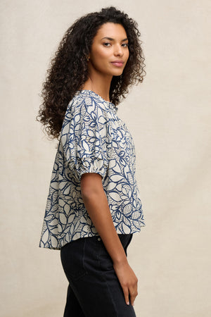 Woman with curly hair stands in profile wearing the THERA TOP by Velvet by Graham & Spencer, a short-sleeve cotton voile blouse with a smocked neckline, paired with dark pants against a neutral background.