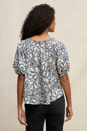 A person with curly hair faces away from the camera against a light background, wearing Velvet by Graham & Spencer's THERA TOP—a blue and white leaf-patterned short-sleeve cotton voile blouse—paired with black jeans.