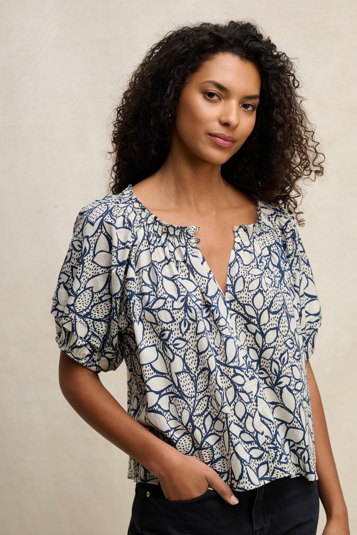 A woman with curly hair stands against a plain background, wearing the Velvet by Graham & Spencer THERA TOP—a cotton voile blouse with short sleeves and a V-neck—with one hand in her pocket.-39539194986689