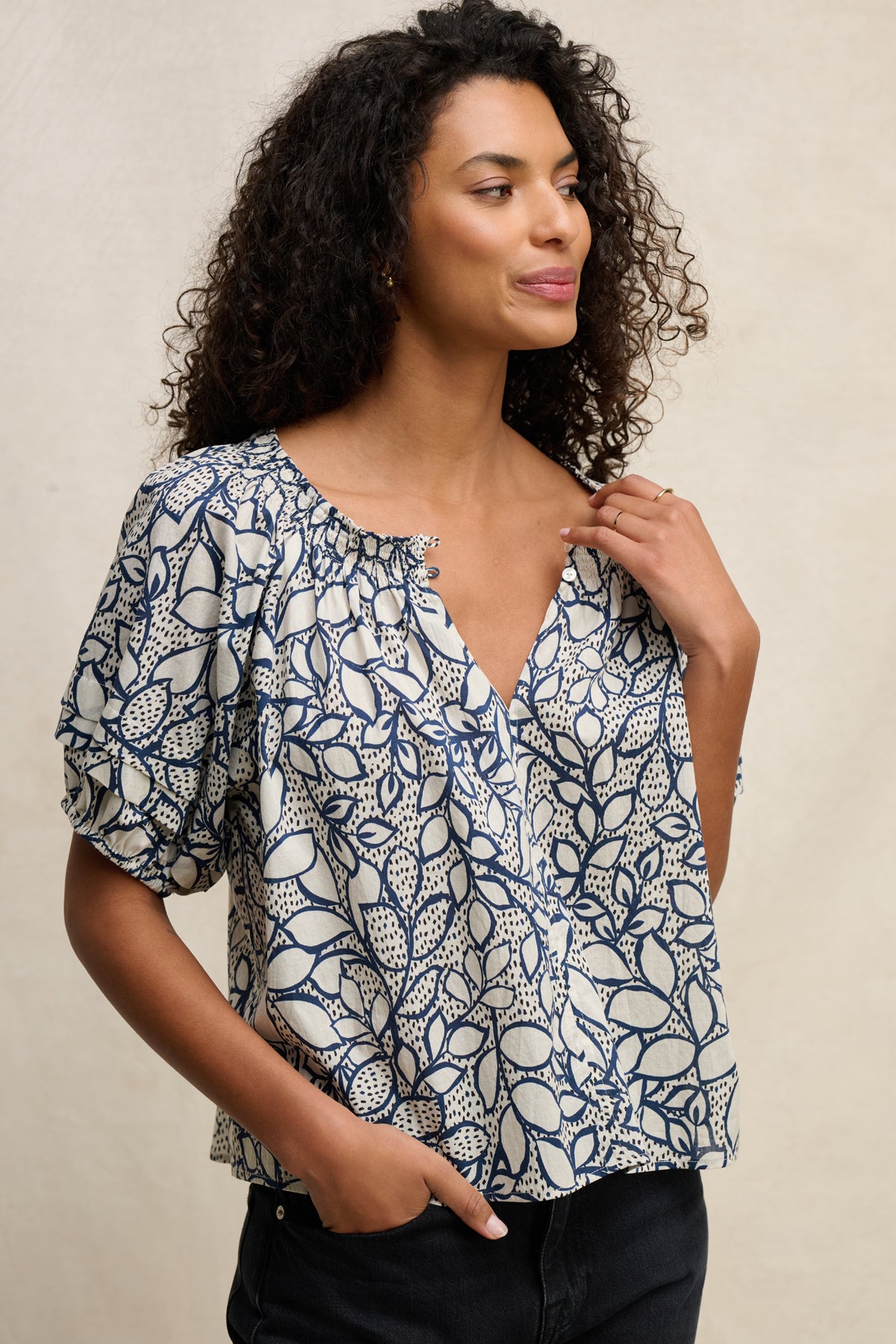 A woman with curly hair wears the THERA TOP by Velvet by Graham & Spencer—a short-sleeve cotton voile blouse with a smocked neckline and blue and white leaf print—paired with black pants against a plain light background.-39539194953921