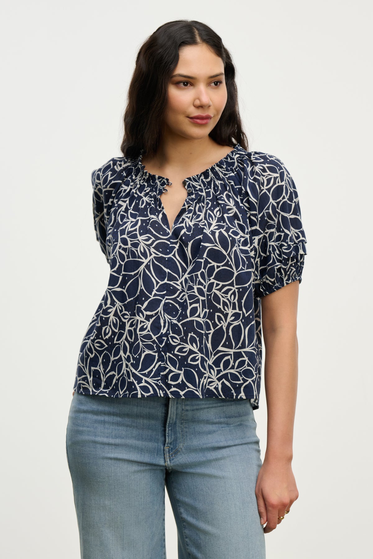 A woman models the THERA TOP by Velvet by Graham & Spencer—a navy blue cotton voile blouse with a white abstract leaf pattern and smocked neckline—paired with light blue jeans, set against a plain light background.-39539194888385