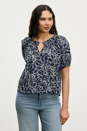 A woman models the THERA TOP by Velvet by Graham & Spencer—a navy blue cotton voile blouse with a white abstract leaf pattern and smocked neckline—paired with light blue jeans, set against a plain light background.