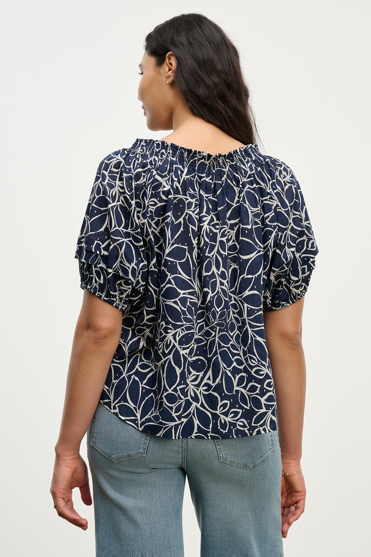 A woman faces away, wearing the THERA TOP by Velvet by Graham & Spencer—a navy cotton voile blouse with a white leaf print, smocked neckline, and short sleeves—paired with light blue jeans.-39560396865729