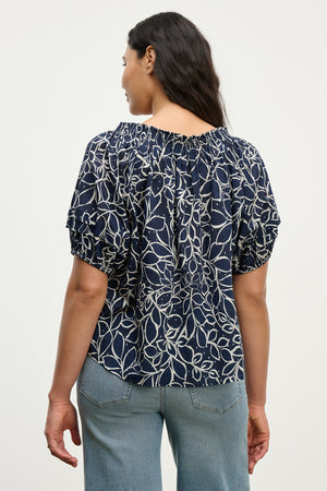 A woman faces away, wearing the THERA TOP by Velvet by Graham & Spencer—a navy cotton voile blouse with a white leaf print, smocked neckline, and short sleeves—paired with light blue jeans.