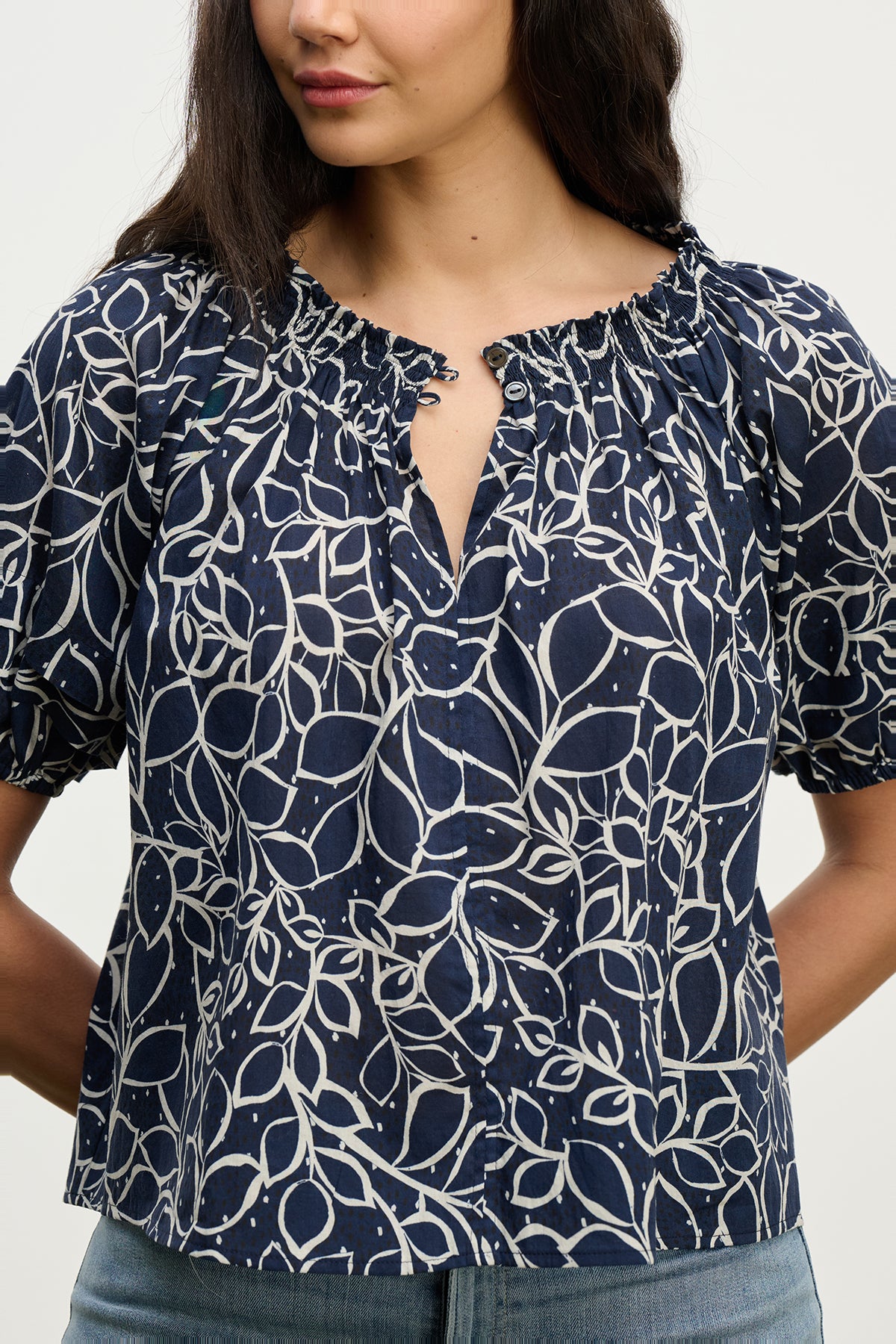 A woman wears the THERA TOP by Velvet by Graham & Spencer—a navy blue cotton voile blouse with a white abstract leaf pattern, smocked buttoned neckline, and short sleeves—paired with light blue jeans.-39560396898497
