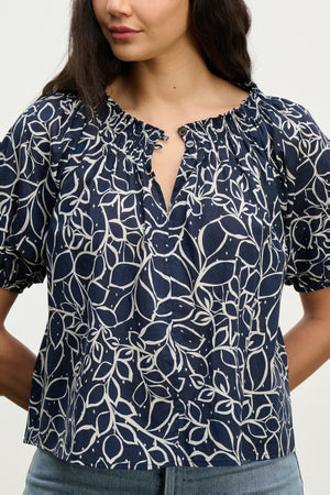 A woman wears the THERA TOP by Velvet by Graham & Spencer—a navy blue cotton voile blouse with a white abstract leaf pattern, smocked buttoned neckline, and short sleeves—paired with light blue jeans.