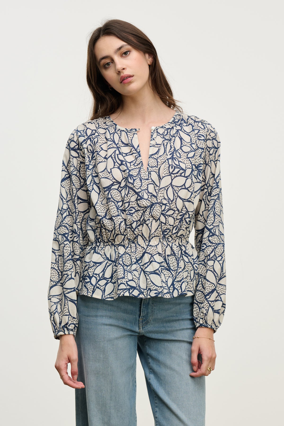 A woman wears the Velvet by Graham & Spencer WYNNE TOP, a long-sleeve cotton voile blouse with a feminine silhouette and navy/beige leaf print, featuring a keyhole closure, paired with light blue jeans.-40227172810945